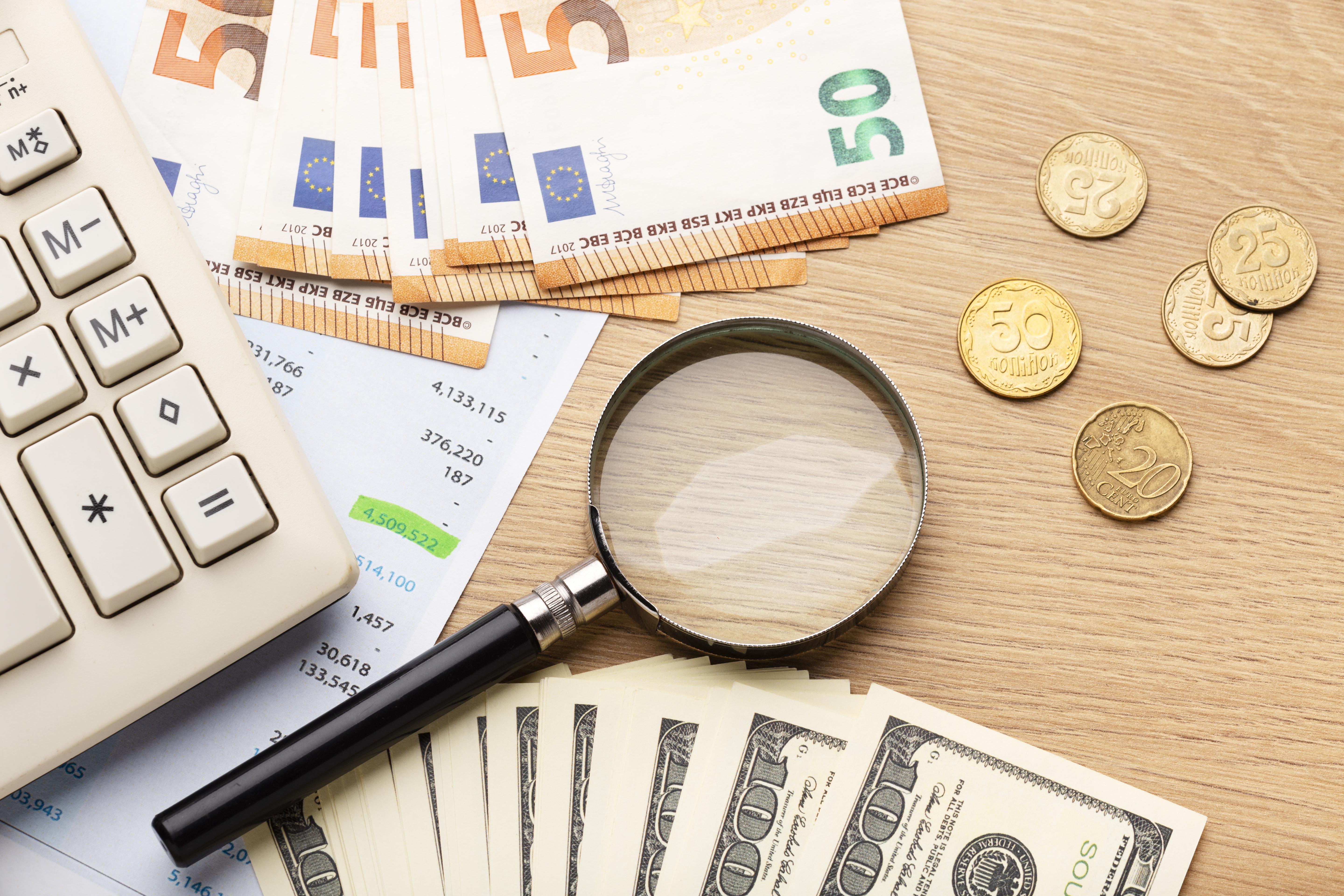 Close-up of financial documents with euro banknotes, U.S. dollar bills, assorted coins, a calculator, and a magnifying glass
