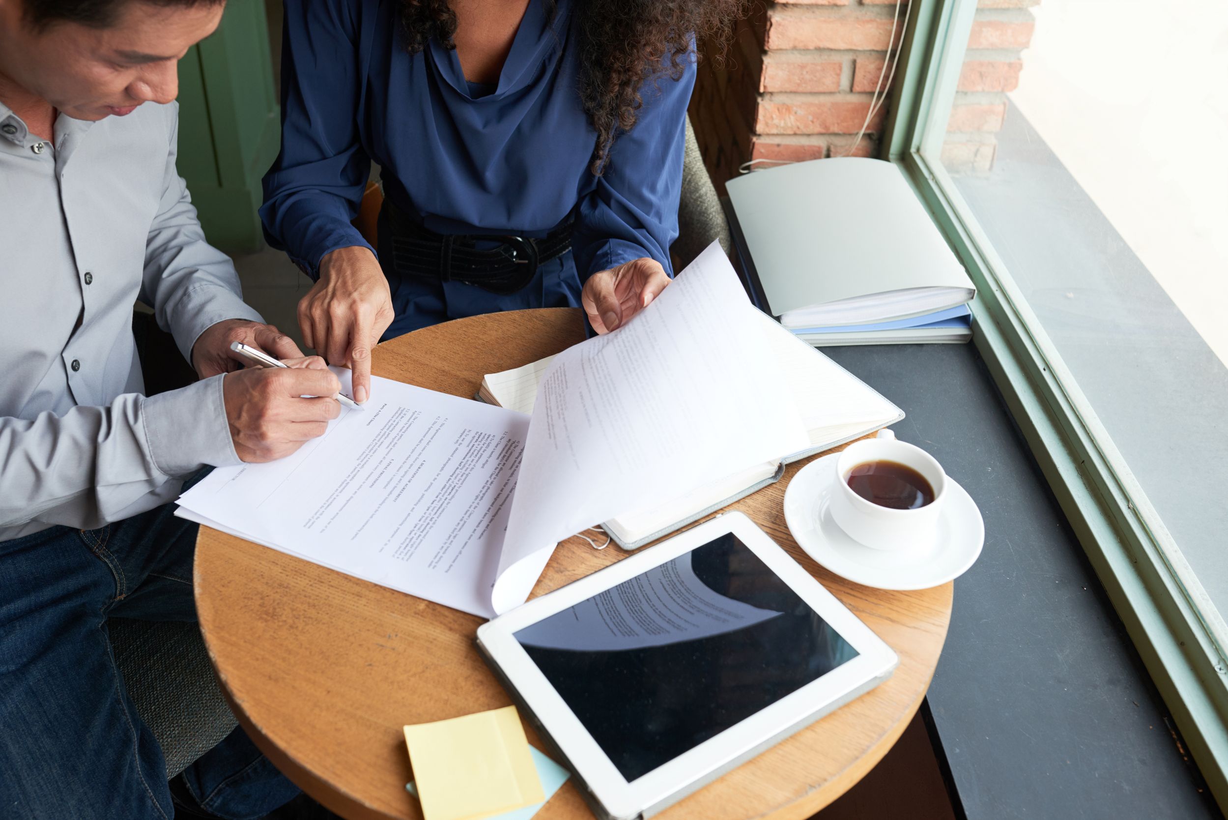 Two colleagues review and sign business documents at a table with a tablet and coffee.